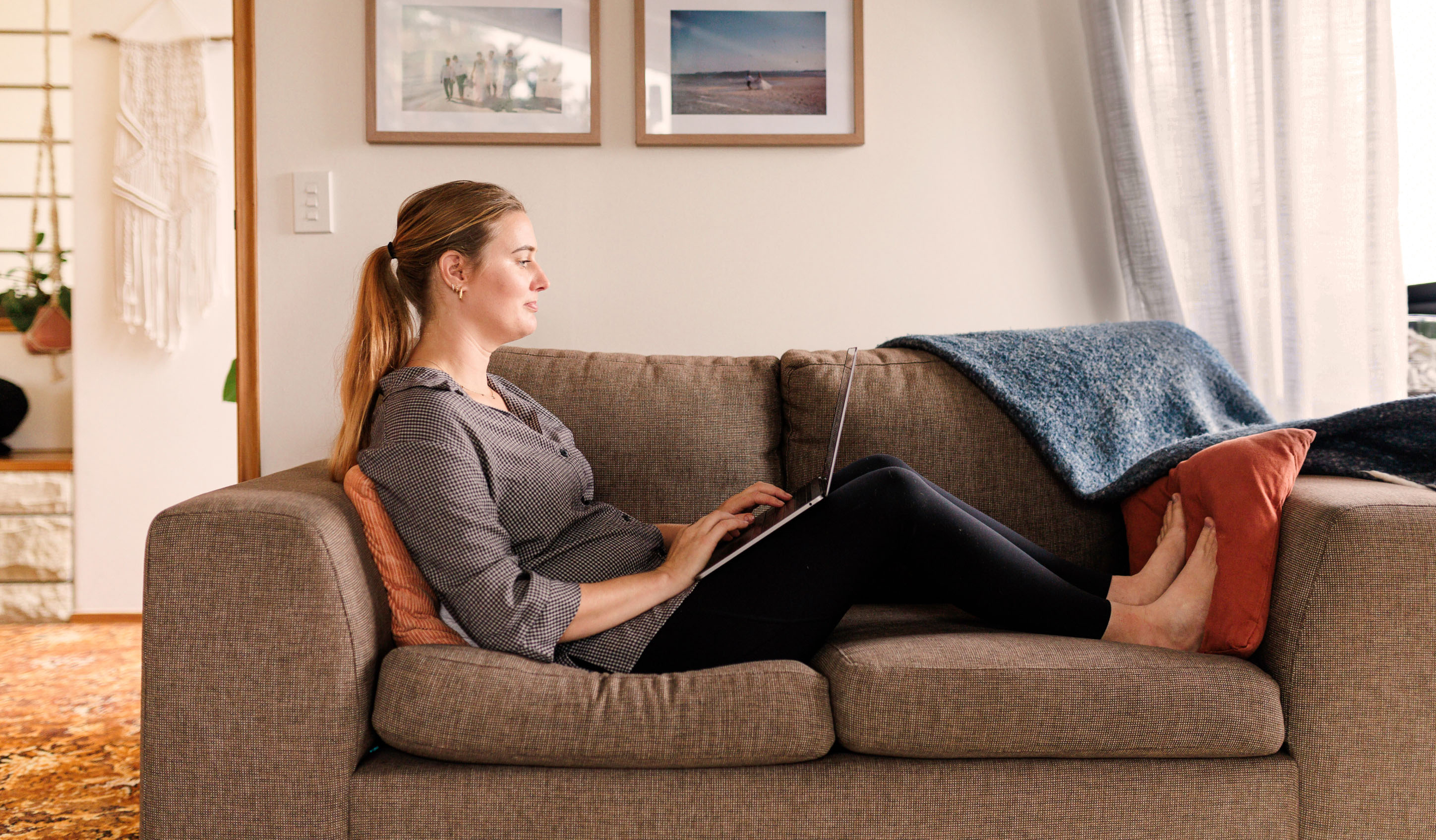 A woman sitting on a couch at home using a laptop, with framed photos on the wall behind her.
